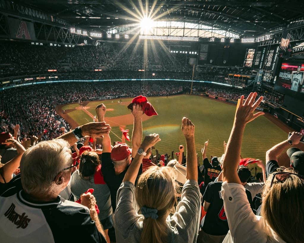 people trying to cheer for a baseball game but the sun is peaking through the lip of the rooftop and is blinding half the crowd